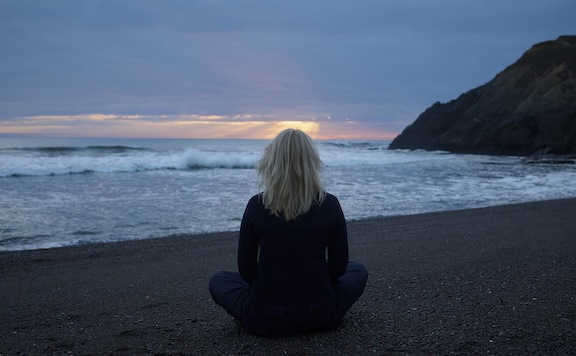 A person with blonde hair sits cross-legged on a sandy beach, deep in meditation as they watch the sunset over the ocean, waves gently rolling and a rocky hill in the background.