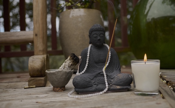 A Buddha statue with a beaded necklace sits on a wooden surface beside a burning candle, incense stick, sage bundle, and a small bowl—creating a peaceful meditation space for mantra practice.