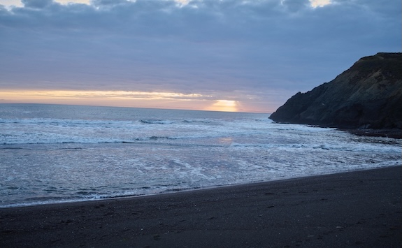 A sandy beach with gentle waves under a cloudy sky at sunset invites meditation, while a rocky cliff rests on the right side of the image.