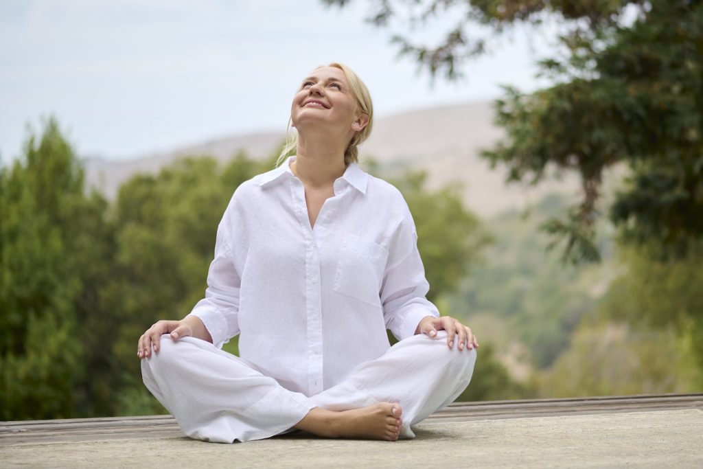 A woman in white clothing sits cross-legged outdoors on a mat, practicing meditation and looking up with a smile, surrounded by trees and greenery.