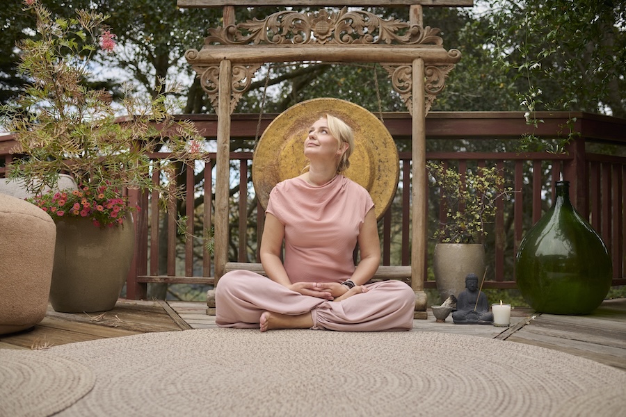 A woman in pink sits cross-legged on a round rug outdoors, surrounded by plants and decor, looking upward with a calm expression as if deep in meditation.