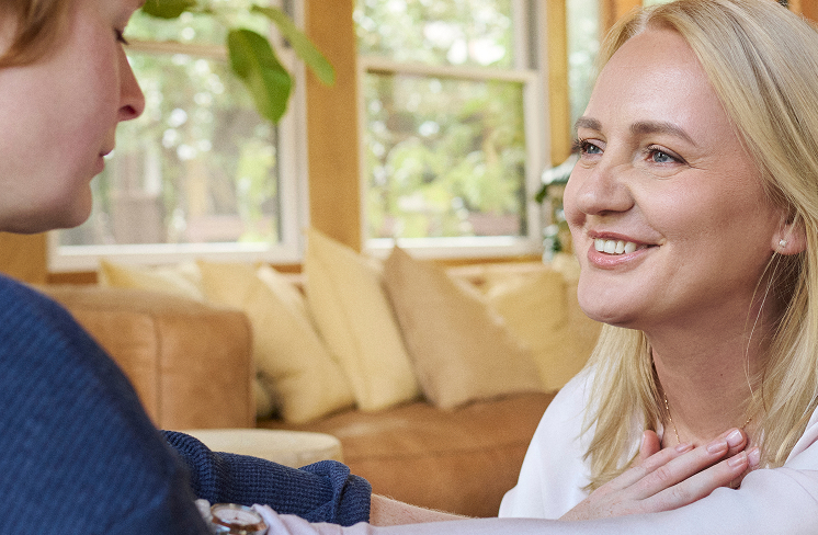 Two people sit indoors; one smiles warmly with hands on chest, as if sharing a calming mantra with the other, who is partially visible. A couch and windows provide a peaceful backdrop.