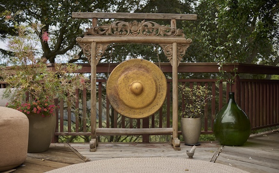 A large brass gong on an ornate wooden stand sits on a wooden deck, surrounded by potted plants and outdoor furniture—an inviting space for meditation or reciting a calming mantra.