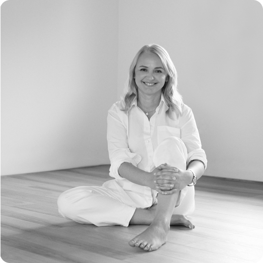 A woman with light hair sits barefoot on a wooden floor, dressed in a light shirt and pants, smiling gently at the camera as she enjoys a moment of meditation in a minimally furnished room.