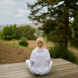A person in white sits cross-legged on a wooden deck, lost in meditation as they face a scenic landscape with trees and hills in the background.