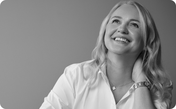 Black and white photo of a smiling woman with long, light hair wearing a white shirt, necklace, and watch, looking upwards with her hand touching her neck—capturing a peaceful moment as if in mantra meditation.