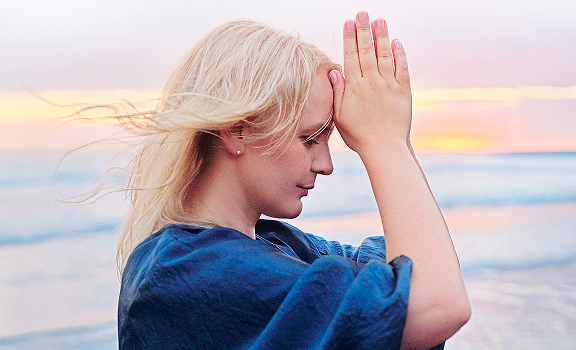 A woman with blonde hair stands near the ocean at sunset, holding her hands together in front of her forehead in a meditation pose, silently reciting a mantra.