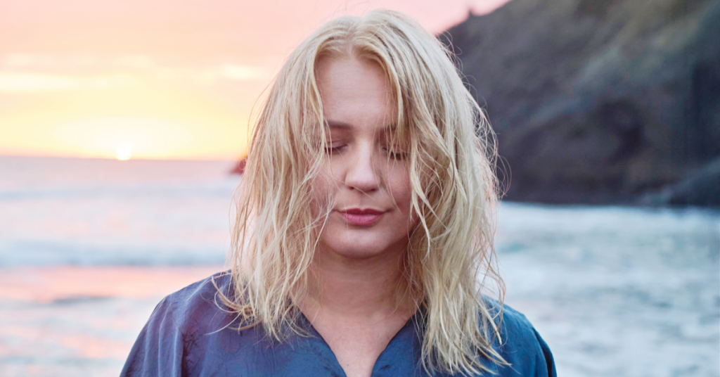 A woman with blonde hair stands near the ocean at sunset, eyes closed in meditation, wearing a blue shirt.