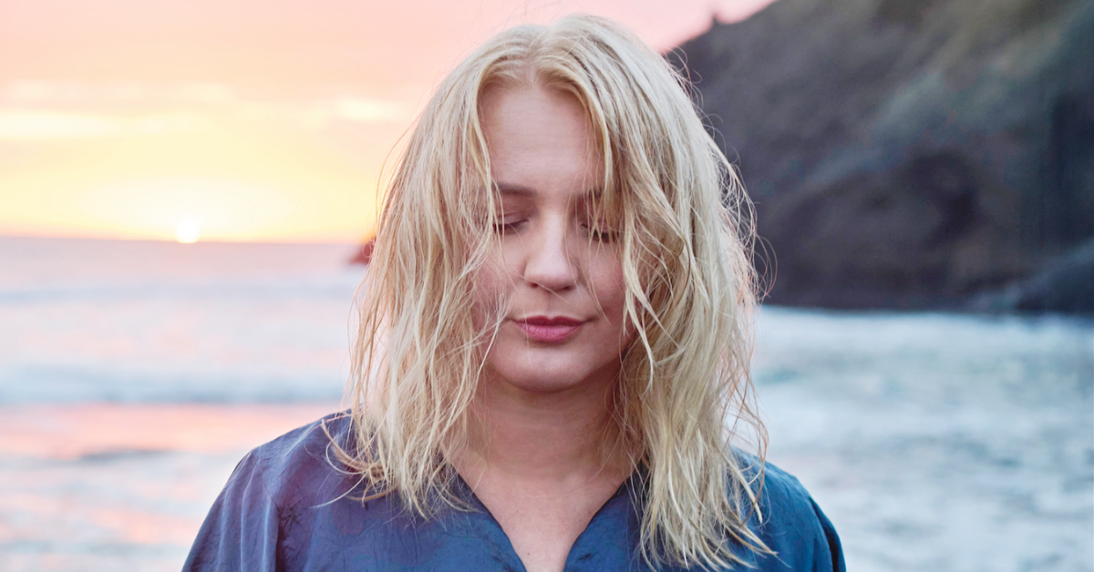 A woman with blonde hair stands near the ocean at sunset, eyes closed in meditation, wearing a blue shirt.