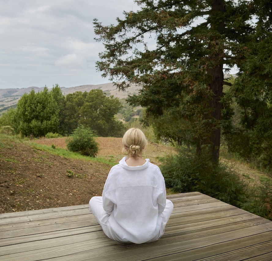 A person in white clothing sits cross-legged on a wooden deck, deep in meditation, facing away and overlooking trees and hills under a cloudy sky.