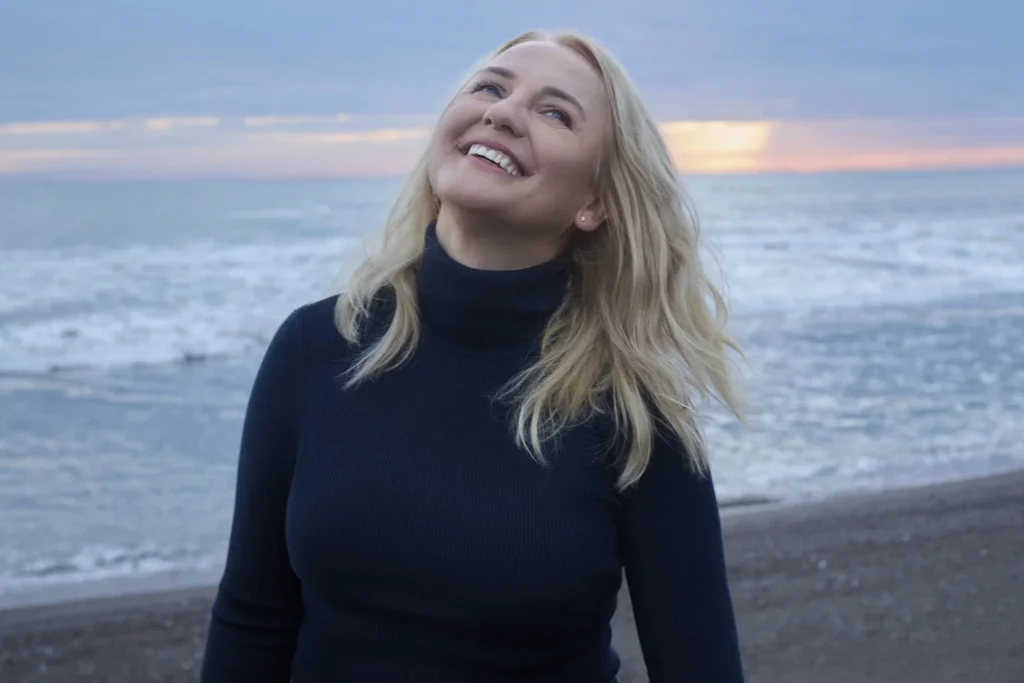 A woman with long blonde hair in a black turtleneck stands on a beach, smiling and looking upwards, as the ocean and sunset create a peaceful backdrop—embracing the calmness of meditation.