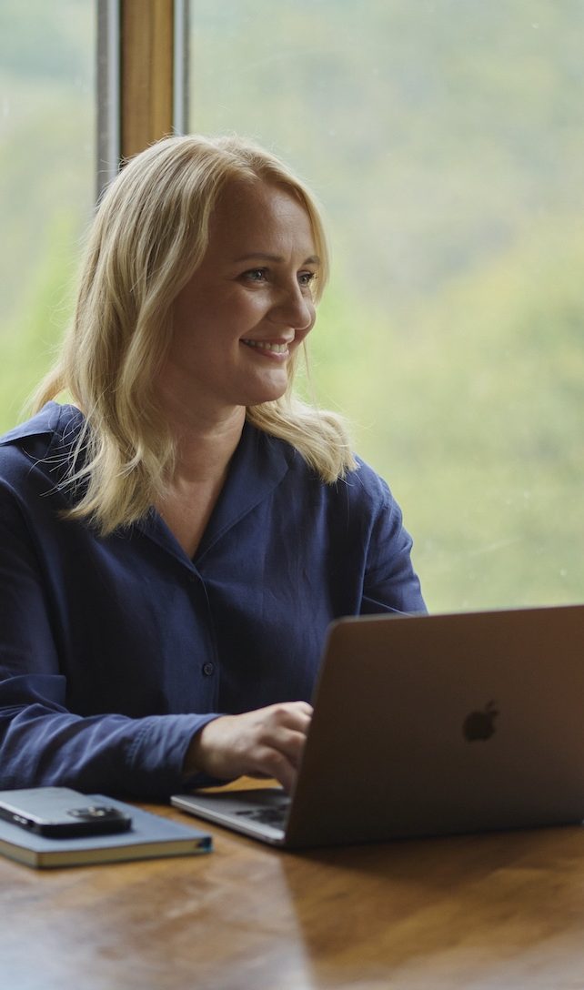 A woman with blonde hair sits at a wooden table, smiling while using a laptop. A notebook and smartphone are on the table beside her, as she enjoys a peaceful moment of meditation. A window with greenery is in the background.