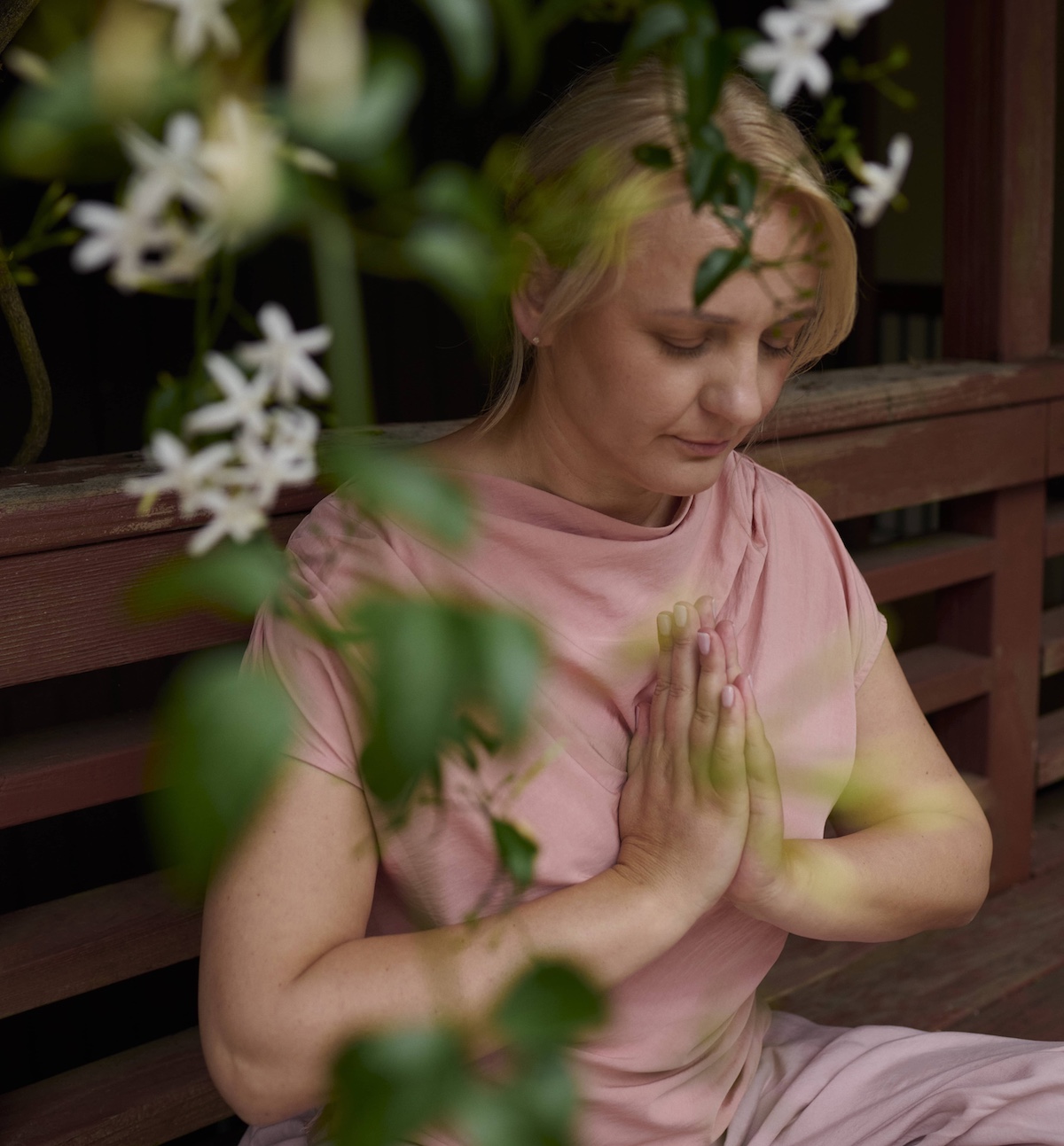A woman in a pink dress sits cross-legged on a wooden porch, hands pressed together in a prayer position, partially obscured by green leaves and white flowers, lost in peaceful meditation.